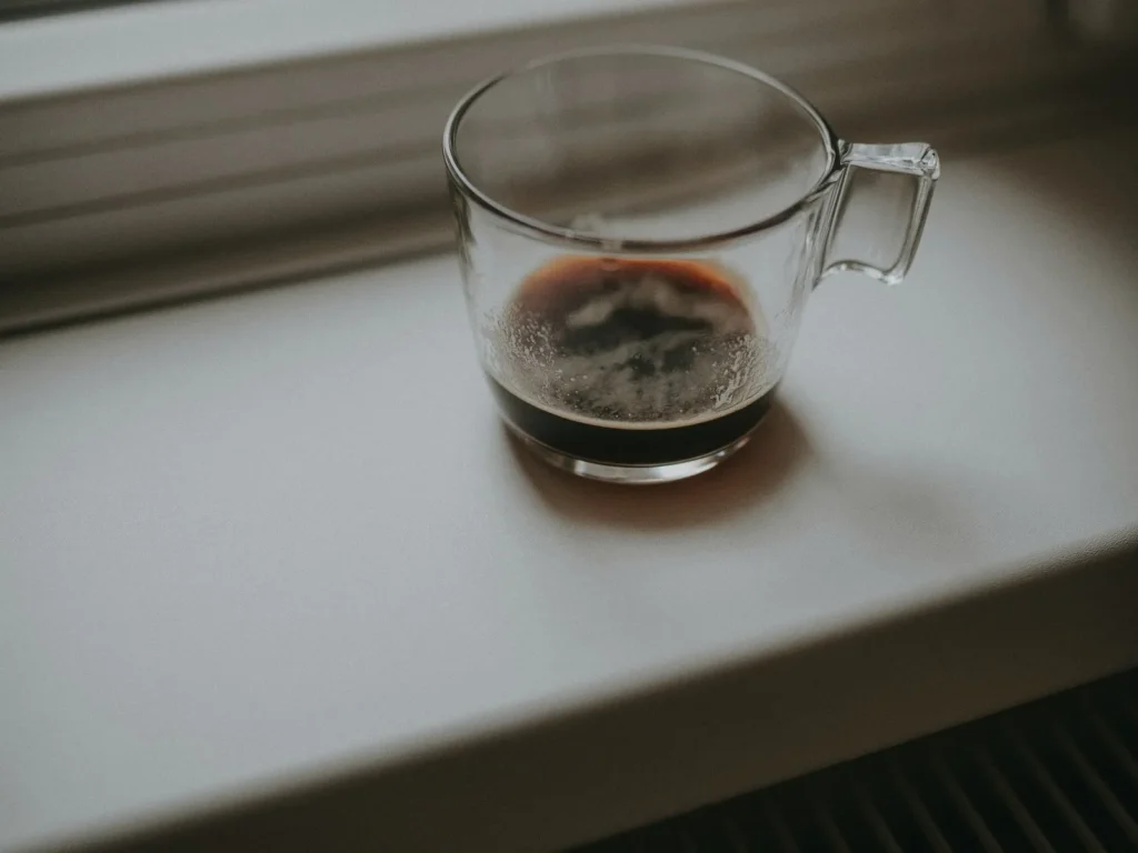 A glass cup with the last of a black coffee on a white windowsill in soft light
